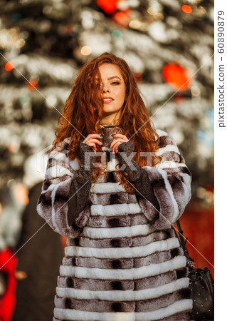 Outdoor portrait of young beautiful redhead happy smiling girl is wearing fur warm hat. Festive Christmas lights on background. 60890879