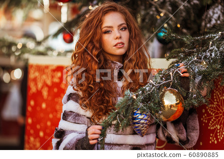 Outdoor portrait of young beautiful redhead happy smiling girl is wearing fur coat near decorated Christmas tree. 60890885