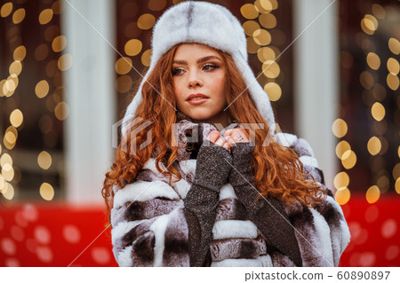 Outdoor portrait of young beautiful redhead happy smiling girl is wearing fur warm hat. Festive Christmas lights on background. Outdoor portrait of young beautiful redhead happy smiling girl is wearing fur warm hat. Festive Christmas lights on background. 60890897