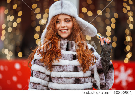 Outdoor portrait of young beautiful redhead happy smiling girl is wearing fur warm hat. Festive Christmas lights on background. 60890904