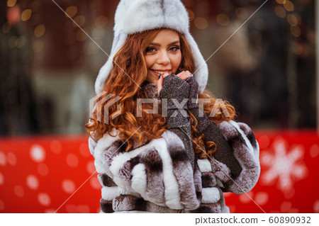 Outdoor portrait of young beautiful redhead happy smiling girl is wearing fur warm hat. Festive Christmas lights on background. Outdoor portrait of young beautiful redhead happy smiling girl is wearing fur warm hat. Festive Christmas lights on background. 60890932
