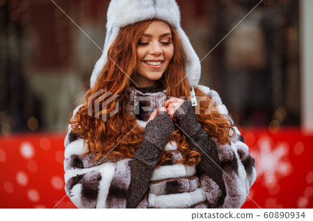 Outdoor portrait of young beautiful redhead happy smiling girl is wearing fur warm hat. Festive Christmas lights on background. 60890934