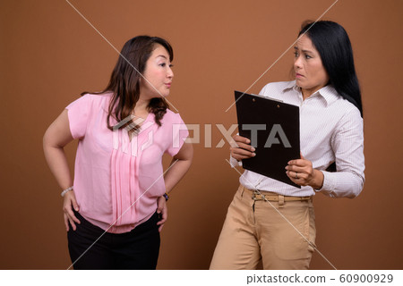 Two mature Asian businesswomen together against brown background Two mature Asian businesswomen together against brown background 60900929