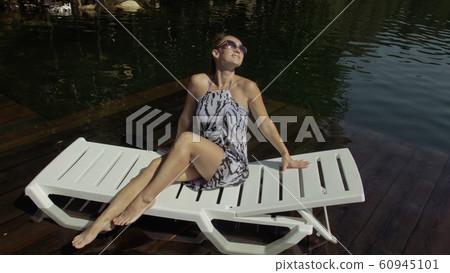 Woman lie on a sunbed in sunglasses and a boho silk shawl. Girl rest on a flood wood underwater pier. The pavement is covered with water in the lake. 60945101
