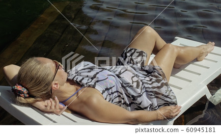 Woman lie on a sunbed in sunglasses and a boho silk shawl. Girl rest on a flood wood underwater pier. The pavement is covered with water in the lake. Woman lie on a sunbed in sunglasses and a boho silk shawl. Girl rest on a flood wood underwater pier. The pavement is covered with water in the lake. 60945181