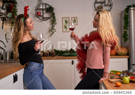 Happy blonde girls celebrating new year with wine glasses in their hands standing in kitchen. Happy blonde girls celebrating new year with wine glasses in their hands standing in kitchen. 60945260