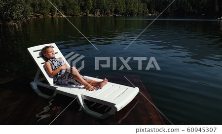 Kid girl lie on a sunbed in sunglasses and a boho silk shawl. Child rest on a flood wood underwater pier. The pavement is covered with water in lake. 60945307