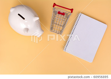 White piggy bank, shopping trolley and notebook on a beige background. View from above. Saving concept. 60956895