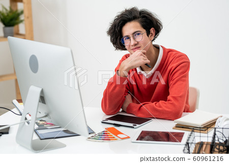 Young successful freelance designer sitting by desk in front of computer monitor 60961216