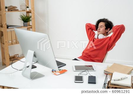 Serene young male freelancer daydreaming by desk with his hands behind head Serene young male freelancer daydreaming by desk with his hands behind head 60961291