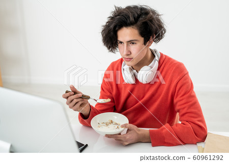 Young male freelancer having muesli with milk and watching online video Young male freelancer having muesli with milk and watching online video 60961292