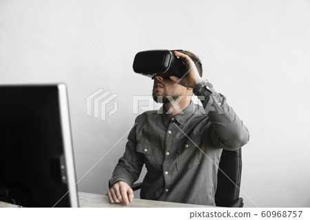 Young bearded man sitting against computer with virtual reality glasses. Modern technologies. The concept of future technology. Young bearded man sitting against computer with virtual reality glasses. Modern technologies. The concept of future technology. 60968757