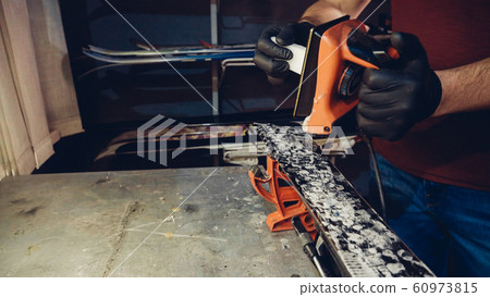 Applying hot wax to ski base with iron. Applying Ski Wax To Ski Base. Ski base tuning. Ski wax iron and wax bars. Ski service and repair. repairman in workshop ski service. Man Waxing Ski In Workshop 60973815