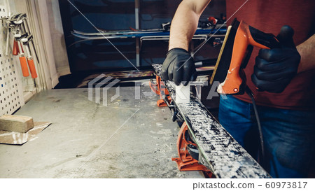 Applying hot wax to ski base with iron. Applying Ski Wax To Ski Base. Ski base tuning. Ski wax iron and wax bars. Ski service and repair. repairman in workshop ski service. Man Waxing Ski In Workshop 60973817