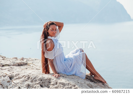 A beautiful woman with long hair in a dress looks out over the sea from a cliff 60987215