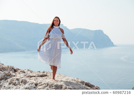 A beautiful woman with long hair in a dress looks out over the sea from a cliff 60987216