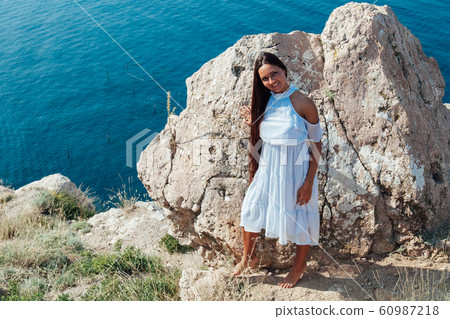 A beautiful woman with long hair in a dress looks out over the sea from a cliff 60987218