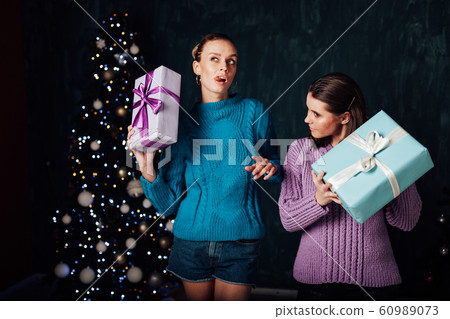 Two beautiful female girlfriends open Christmas gifts at the Christmas tree for the New Year holiday Two beautiful female girlfriends open Christmas gifts at the Christmas tree for the New Year holiday 60989073