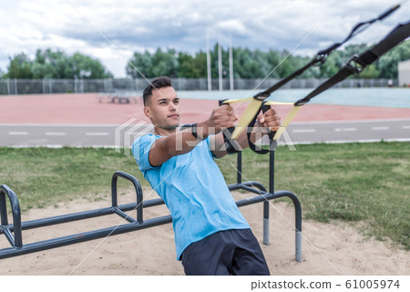 Young sportsman, male athlete, pulls himself on hinges, straps for chest muscles, in summer day in the city, workout fitness, active lifestyle of modern youth, sportswear. Motivation for life. Young sportsman, male athlete, pulls himself on hinges, straps for chest muscles, in summer day in the city, workout fitness, active lifestyle of modern youth, sportswear. Motivation for life. 61005974