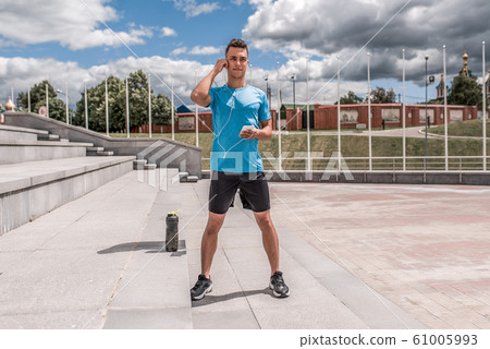 Young sportsman, male athlete, listens music with headphones, summer city, workout with fitness training, active lifestyle, sportswear t-shirt shorts sneakers. Motivation life. Free space copy text. Young sportsman, male athlete, listens music with headphones, summer city, workout with fitness training, active lifestyle, sportswear t-shirt shorts sneakers. Motivation life. Free space copy text. 61005993