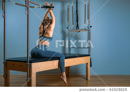 Young girl doing pilates exercises with a reformer bed. Beautiful slim fitness trainer on reformer gray background, low key, art light. Fitness concept 61029829