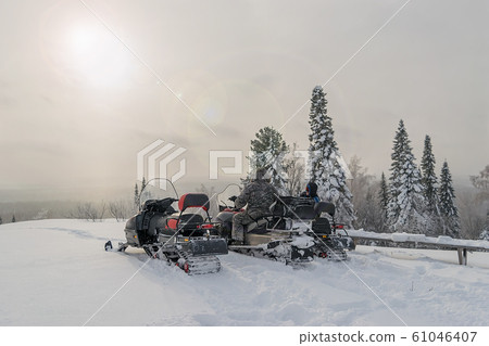 people sit and talk on snowmobiles at the top of the mountain against the background of the forest and the sun glare in the cold winter 61046407