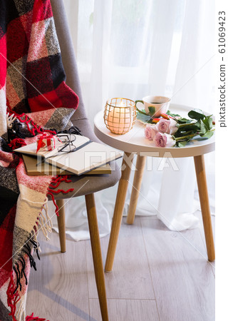 A candle with a notebook on a table with pink roses in a box near a window with a chair, a plaid and a book A candle with a notebook on a table with pink roses in a box near a window with a chair, a plaid and a book 61069423