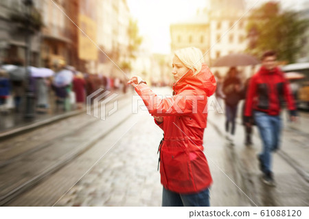Time concept. Girl looking on watch and waiting friend on the street. Portrait of pretty smiling young woman in red bright raincoat. Sunny raining day in city. Image with yellow flare 61088120