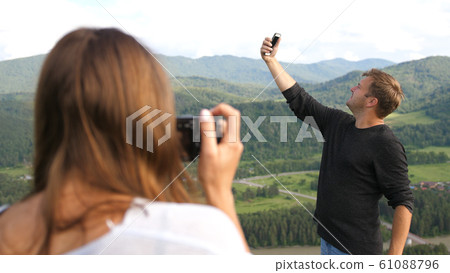 Tourists couple enjoying the nature in mountains, young woman taking photos of man that takes selfie on the phone. 61088796