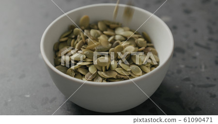 closeup dried pumpkin seeds falling into white bowl on terrazzo countertop 61090471