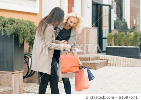 Two young smiling women on a city street with shopping bags, sunny autumn day, golden hour Two young smiling women on a city street with shopping bags, sunny autumn day, golden hour 61097671