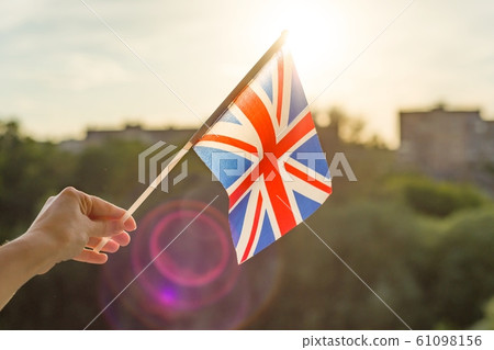 Hand holding Great Britainin flag an open window. Background blue sky, silhouette of the city, sunset Hand holding Great Britainin flag an open window. Background blue sky, silhouette of the city, sunset 61098156