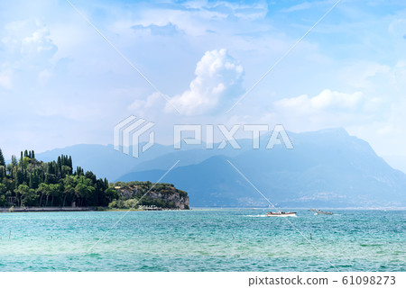 Panoramic view against bright sun rays above the rocks on beautiful Garda lake shore in Riva del Garda city in Lombardy, Italy, surrounded by high dolomite mountains 61098273