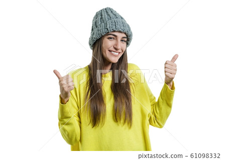 Young beautiful woman in yellow sweater and gray big loop knitted beanie hat, showing sign ok thumbs up, looking at the camera on white isolated background 61098332