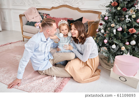 Loving family. Mom and dad hugging little daughter . Parents and baby child having fun near Christmas tree and white fireplace indoors. Merry Christmas and Happy New Year. Cheerful pretty people. Loving family. Mom and dad hugging little daughter . Parents and baby child having fun near Christmas tree and white fireplace indoors. Merry Christmas and Happy New Year. Cheerful pretty people. 61109893