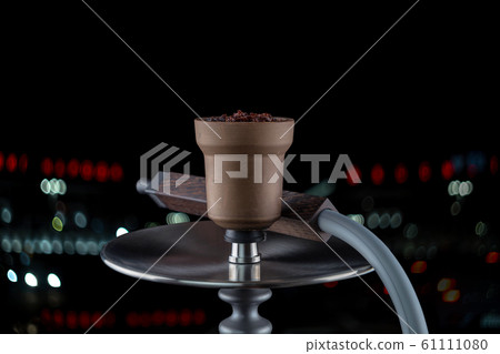 Clay bowl with shisha on a modern Smoking hookah with a mouthpiece of beautiful wood on a dark background with colored bokeh. Copy space. Clay bowl with shisha on a modern Smoking hookah with a mouthpiece of beautiful wood on a dark background with colored bokeh. Copy space. 61111080