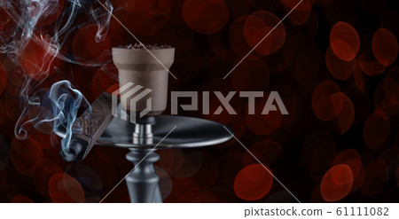 Hookah, from the mouthpiece of which curls a beautiful smoke on a dark beautiful red bokeh background. Hookah, from the mouthpiece of which curls a beautiful smoke on a dark beautiful red bokeh background. 61111082