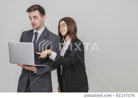 Handsome man standing, working with laptop. Woman is pointing from man's behind at something in the laptop, interfering him. Man is annoyed because of this. Handsome man standing, working with laptop. Woman is pointing from man's behind at something in the laptop, interfering him. Man is annoyed because of this. 61137376