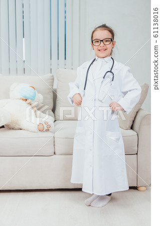 Pretty confident little girl wearing doctor coat, stethoscope and glasses, standing, holding hands akimbo, looking at the camera, smiling. 61138016