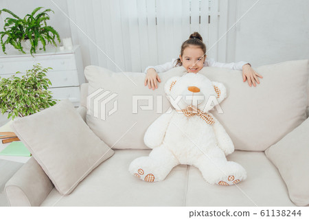Big white soft bear is on the sofa. Girl is standing behind the sofa, leaning over its back, looking at the camera, smiling pleasantly. Big white soft bear is on the sofa. Girl is standing behind the sofa, leaning over its back, looking at the camera, smiling pleasantly. 61138244