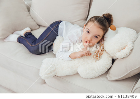 Nice little girl is looking at the camera, showing a silence sign while lying on her big white teddy-bear. Full-length. Nice little girl is looking at the camera, showing a silence sign while lying on her big white teddy-bear. Full-length. 61138409
