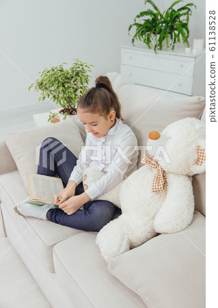 Cute little girl sitting on the sofa with teddy-bear near her, reading fairy-tale. Cute little girl sitting on the sofa with teddy-bear near her, reading fairy-tale. 61138528