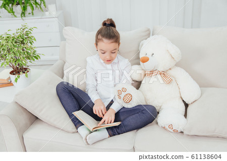 Cute little girl sitting on the sofa with teddy-bear near her, reading fairy-tale. Cute little girl sitting on the sofa with teddy-bear near her, reading fairy-tale. 61138604