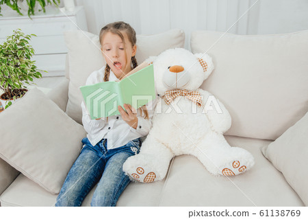 Cute little girl sitting on the sofa with teddy-bear near her, reading fairy-tale. Cute little girl sitting on the sofa with teddy-bear near her, reading fairy-tale. 61138769
