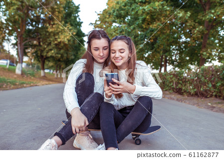 2 girls girlfriends 12-15 years old, happy sit smile, relax skateboard, city, rejoice and have fun. In hands of smartphone, watching videos online app social networks, casual clothes, jeans, sweater. 61162877