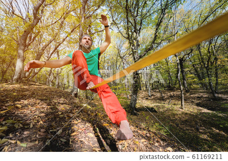 Wide angle male tightrope walker balancing while sitting barefoot on slackline in autumn forest. The concept of outdoor sports and active life of people aged 61169211