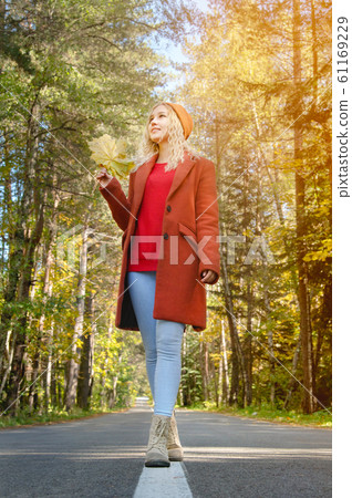 Attractive happy woman in a red coat smiling joyful and blissful holding autumn leaves outside in her hand in a colorful autumn forest walks on asphalt road. 61169229