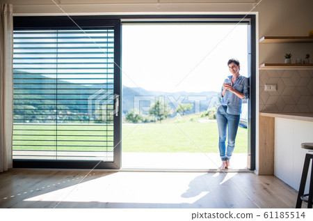 Front view of young woman with coffee standing by patio door at home. Front view of young woman with coffee standing by patio door at home. 61185514