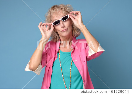 Senior woman wearing stylish sunglasses and smiling at camera on blue wall. 61229044