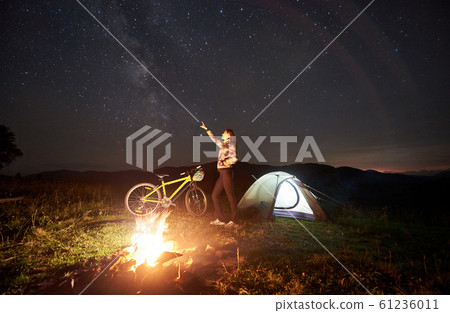 Woman resting at night camping near campfire, tourist tent, bicycle under evening sky full of stars Woman resting at night camping near campfire, tourist tent, bicycle under evening sky full of stars 61236011